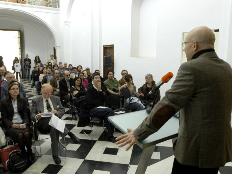 Un hombre en traje habla frente a una audiencia en un salón con pisos negros y blancos. La audiencia está sentada en filas, mirando al hablante.