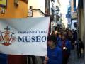 Mujer sostiene un cartel blanco con el nombre "Hermandad del Museo" y una cruz roja, mientras camina por un callejón en una ciudad.