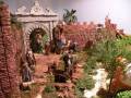 Una escena navideña con figuras de madera y piedra, incluyendo un arco, una palmera y personajes vestidos con ropas antiguas. La escena está rodeada por vegetación artificial y una estructura de madera con un asiento.