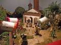 Una escena navideña con figuras de madera y adornos navideños en un entorno rural. Hay dos carros con flores, una casa con chimenea y un árbol en el fondo. La imagen parece ser parte de una escena navideña o un set de decoración.