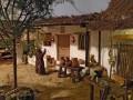 Escena colonial con una casa blanca, un hombre vestido de rojo y una mesa con objetos.