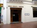El Círculo Mercantil e Industrial es un edificio histórico ubicado en la calle, con una entrada principal de madera y un letrero que indica su nombre. La fachada es blanca con detalles decorativos en la parte superior.