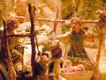 Un escenario de la vida cotidiana en un pueblo medieval, con figuras humanas en pie, sosteniendo objetos y vestidos con colores vibrantes, rodeadas de un entorno que incluye árboles y estructuras de madera.