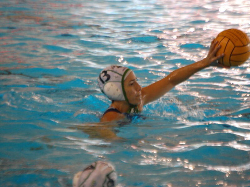 Una jugadora de waterpolo con un casco blanco y verde, preparándose para lanzar una pelota de waterpolo en una piscina azul.