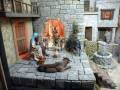 Escena navideña con personajes bíblicos en un entorno medieval. Un burro y una oveja descansan, mientras una persona sostiene un cordero. Detalles como la fuente de agua y las casas de piedra completan el paisaje.