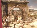 Un pequeño pueblo medieval con una arquitectura de piedra y un arco en el centro. Hay personas vestidas con túnicas antiguas, ovejas y un ganadero en el centro. El paisaje al fondo muestra montañas y cielo azul.