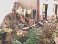 Una escena de una casa colonial con un establo adornado con plantas y flores, incluyendo una figura de un caballo y un niño. La casa tiene paredes rojas con ventanas y puertas, y hay una figura de un niño vestido con ropa de época.