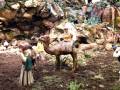 Una escena navideña con figuras de madera en un paisaje rocoso. Una figura femenina monta un camello, mientras una mujer con sombrero y vestido largo se prepara para subir al animal. La escena está rodeada de rocas y vegetación, con un cielo nublado.