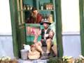 Un miniatura de un hombre sentado en una silla dentro de una puerta abierta, con una cesta y flores alrededor.