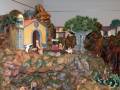Escena navideña con figuras de belenes en un paisaje rocoso, incluyendo una iglesia azul y amarilla con un arco, figuras humanas y animales, árboles y rocas.
