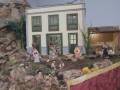 Un pequeño pueblo con una casa blanca y azul, rodeada de vegetación y figuras de personas en miniatura, creando una escena idílica y colorida.