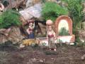 Un entorno natural con rocas y plantas, dos figuras femeninas en trajes tradicionales cargando recipientes, un arco de piedra y una fuente.