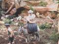 Una niña monta un burro en una escena rural, rodeada de vegetación y rocas.