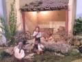 Un entorno de pesebre navideño con figuras humanas y animales, incluyendo un hombre con un animal en su regazo y un hombre con una cesta en el suelo, todo en un entorno con una estructura de techo rojo y vegetación alrededor.