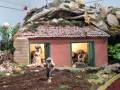 Un niño lleva una cesta en la mano mientras camina hacia un pequeño edificio de ladrillos con una mujer y un niño dentro. La escena parece ser parte de un diorama o escultura, con detalles como una terraza y vegetación alrededor.