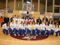 Equipo femenino de karate posando en el piso del gimnasio, con medallas y uniformes blancos y azules.