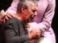 Un hombre en una camisa negra está siendo sujetado por el cuello por otro hombre con una camisa rosa. Ambos parecen estar en un escenario teatral, posiblemente durante una actuación dramática.