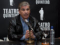 Un hombre habla en un micrófono frente a una pared con el texto "Teatro Quintero" y su imagen. A su lado, hay una botella de agua y un vaso vacío sobre la mesa.