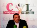 Persona hablando en rueda de prensa con micros y fondo con logo "Compromiso con Lora"