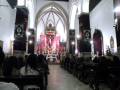 Interior de una iglesia con arcos góticos, vitrales y un altar central. Personas sentadas en bancos de madera.