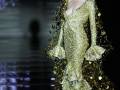 Una modelo desfilando en la noche, luciendo un vestido dorado con detalles brillantes y una cola larga que resplandece en el fondo oscuro.
