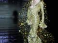 Una modelo con un vestido dorado brillante y detalles de hojas encaja en una pista de desfile nocturna con fuegos artificiales al fondo.