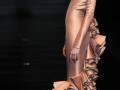 Modela con vestido de satén dorado y falda volante, complementada con sombrero y joyería, en desfile de moda.
