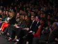 Una audiencia en un evento formal, con personas sentadas y algunas levantando sus teléfonos. La imagen muestra una variedad de estilos de vestimenta, desde trajes formales hasta prendas más informales. La atención se centra en la audiencia, capturando un momento de expectación o interés.