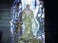 Una modelo desfilando en un vestido dorado con detalles brillantes, posiblemente en una pasarela de moda.