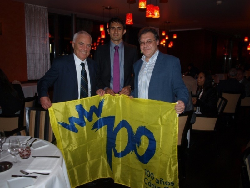 Tres personas sostienen una bandera amarilla con letras azules que dicen '100 años' en un ambiente formal con mesas servidas y luces colgantes rojas.