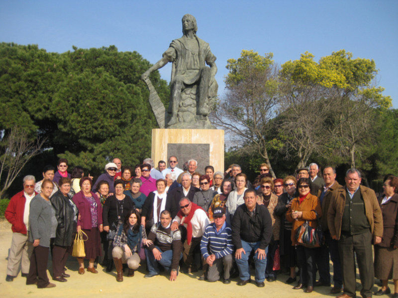 Grande grupo de personas posando frente a una estatua al aire libre, rodeadas por árboles verdes.