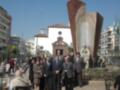 Un grupo de personas en trajes se encuentran en un parque público, frente a una escultura monumental. En el fondo, se puede ver una torre y edificios de varios pisos bajo un cielo azul.