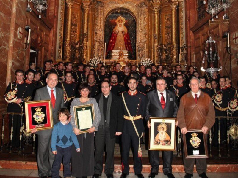 Una familia posa con sus premios en un ambiente histórico, rodeada de uniformes militares y una imagen religiosa en el fondo.