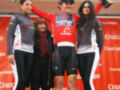 Ciclista con camiseta roja y trofeo en el podio con mujeres vestidas de ciclismo y flores.