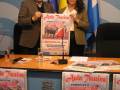 Un hombre y una mujer sostienen un cartel de la Escuela Municipal de Tauromaquia de Jerez, con banderas española y europea al fondo.