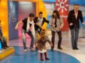 Una niña pequeña y tres hombres adultos están en una escena de televisión, posiblemente un programa infantil. La niña está caminando con los hombres a su lado, todos parecen estar en movimiento. La escena tiene un fondo de colores brillantes y los hombres están vestidos formalmente, uno con una chaqueta gris.