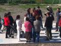 Una multitud de personas se reúne en un área abierta, posiblemente para una reunión o evento. Algunos están en grupos, mientras que otros se desplazan por el lugar. La escena parece ser informal y social, con algunos individuos en trajes formales, lo que sugiere una combinación de eventos sociales y oficiales. En el fondo, se pueden ver árboles y una estructura de metal, lo que indica un entorno urbano o semi-urbano.