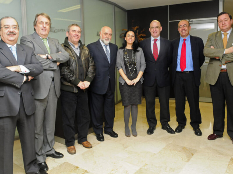 Grupo de personas posando en oficina moderna con paredes de cristal y azulejos en el piso. Todos están vestidos formalmente, con trajes, chaquetas y camisas. La imagen muestra una reunión o presentación en un entorno profesional.