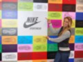Una mujer posa frente a un muro de colores con diferentes siluetas de zapatillas Nike. La imagen destaca el logotipo Nike y la palabra "Sportswear" en la parte superior.