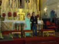Un matrimonio en una iglesia de estilo barroco, con la novia en un vestido azul y el novio en una camisa roja, frente a un altar dorado con velas y flores.