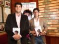 Dos hombres sostienen libros con una imagen de un hombre en la portada. El fondo muestra una pared con carteles y fotos.