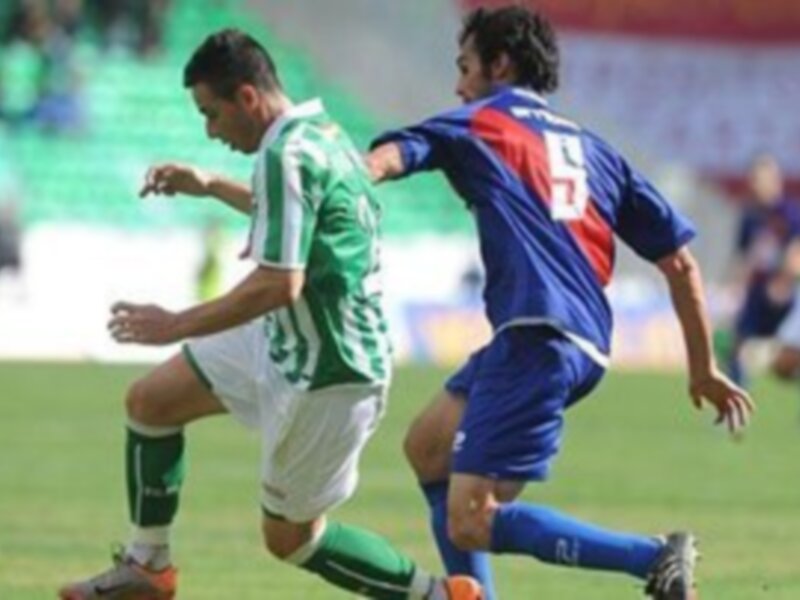 Dos jugadores de fútbol en acción, uno con camiseta verde y otro con camiseta azul, en un campo de juego verde.