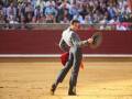 Un torero en acción, con un traje tradicional y un capote rojo, en una plaza de toros. El fondo muestra a la multitud observando el espectáculo.