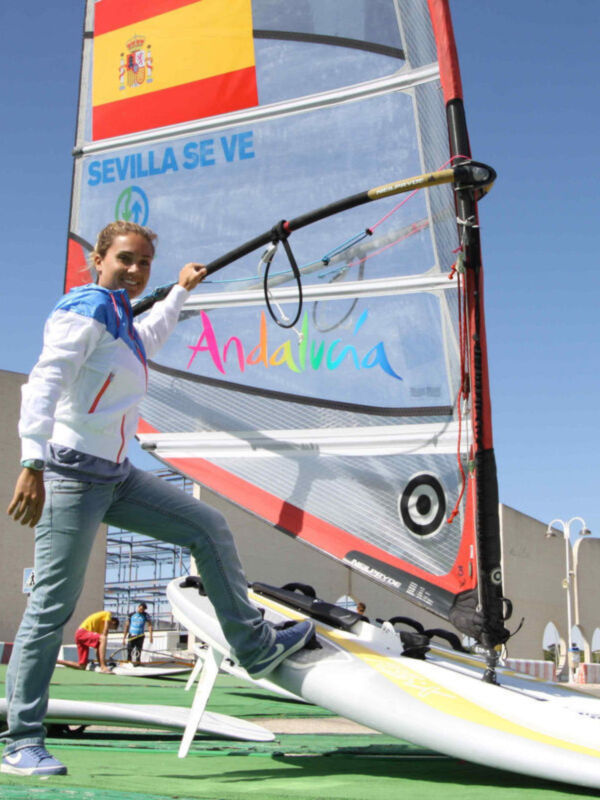 Una persona disfrutando del windsurfing en Sevilla, Andalucía. La vela muestra el nombre de la región y el cielo azul de fondo.