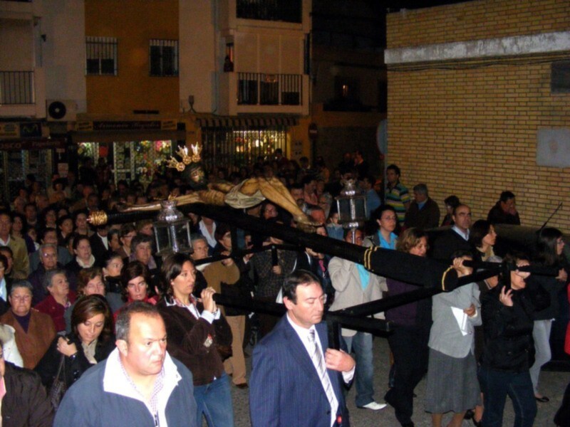 Fotografía de una procesión religiosa en una calle de una ciudad, con una imagen venerada en una mano de un hombre y una multitud de personas observando la procesión.
