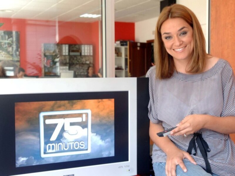 A una mujer sonriendo mientras sostiene un teléfono móvil. En el fondo, se puede ver una pantalla de televisión con la imagen de "75 minutos" y un fondo de cielo nublado. La pared del fondo es roja, y hay una puerta de madera a la derecha.