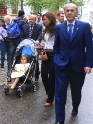 Una familia camina por la calle, con una niña en un cochecito y dos adultos. La imagen muestra a una mujer con una chaqueta blanca, un hombre en traje azul y una niña con zapatos de tacón. La escena parece ser en un día lluvioso, dado el aspecto mojado de la calle.