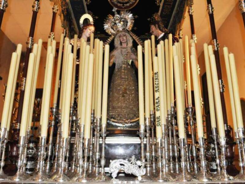Una estatua religiosa en un altar, rodeada de velas encendidas. La estatua parece ser una Virgen María, con un manto dorado y joyas. A su lado, hay dos personas vestidas formalmente, posiblemente fieles o sacerdotes. El ambiente es de una iglesia o templo, con una arquitectura clásica y luces doradas que iluminan la escena.