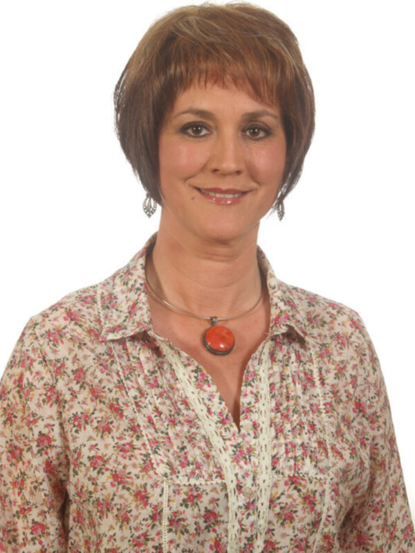 Una mujer con cabello corto y ondulado, lleva una camisa de flores beige con detalles en rosa. Lleva un collar con un colgante redondo de color naranja.