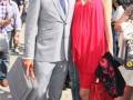 Un hombre en traje gris y corbata azul posa junto a una mujer vestida de rojo, ambos con maletas. En el fondo, se observan personas y un autobús en una calle.